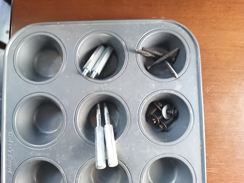 Hardware from disassembly organized in a muffin tin — hex standoffs and Phillips screws in separate cups