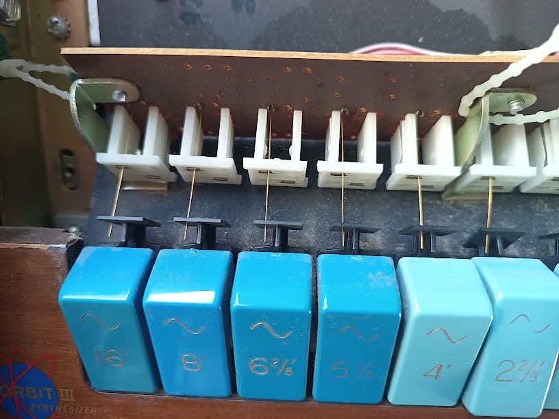 Stop tabs viewed from behind — white plastic plungers and fine gold contact wires visible, with foot-footage markings on the blue tab faces
